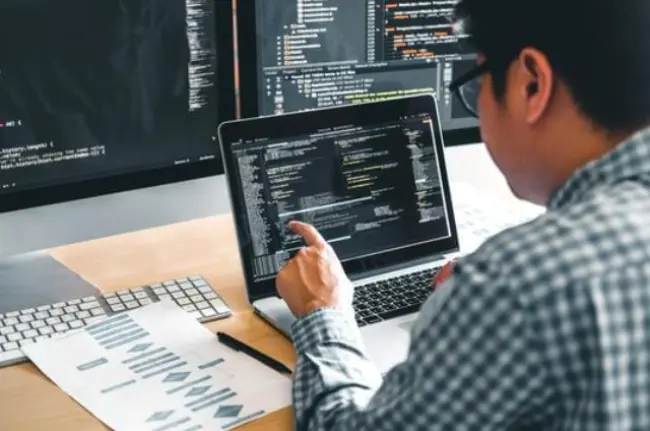 A person in a checked shirt works on coding at a laptop surrounded by multiple screens displaying lines of code and design drafts.