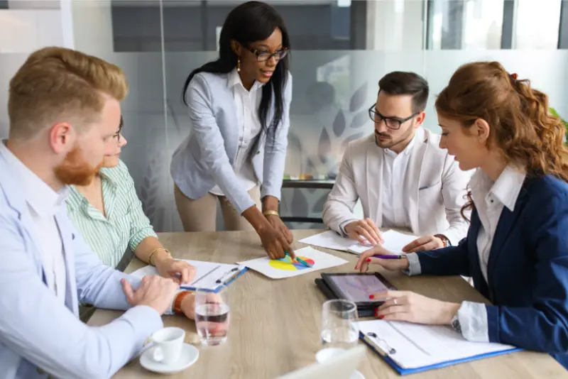 A diverse group of professionals collaborates around a conference table, analyzing charts and discussing strategies during a meeting.