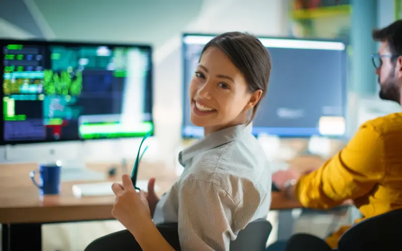 Two individuals focus on multiple computer screens displaying data and graphs in a modern office setting, with a coffee cup nearby.