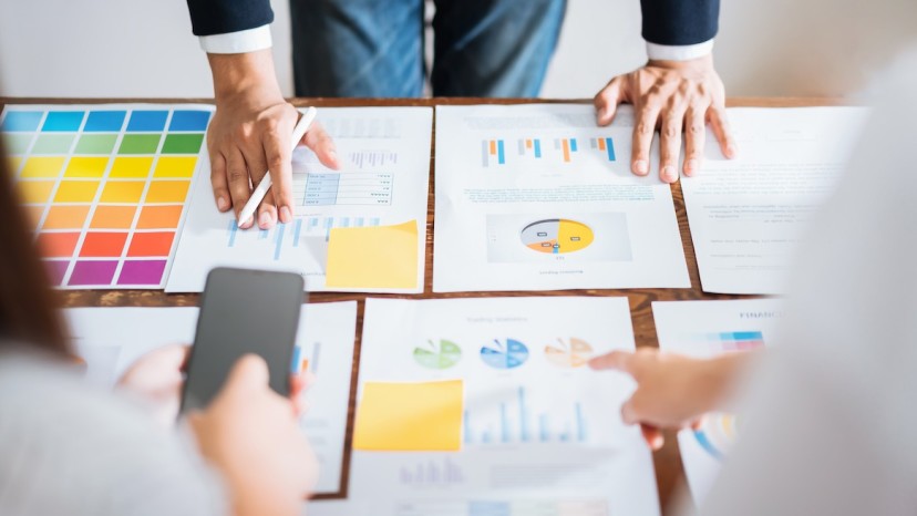 A table covered with colorful charts and graphs, with hands pointing and holding a smartphone, indicating collaboration on data analysis.
