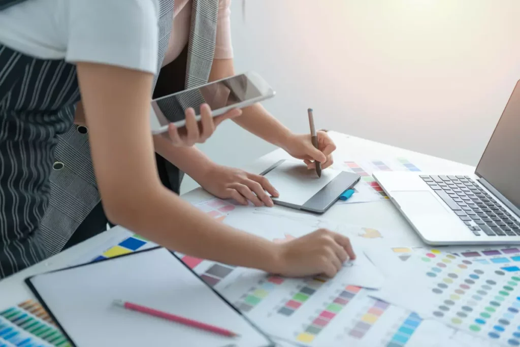Two designers collaborate at a desk, one sketching on a tablet while the other holds a smartphone, surrounded by color swatches and sketches.