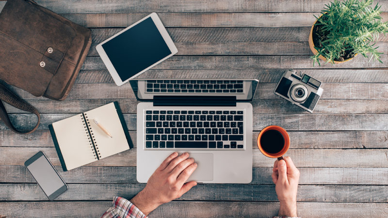 A top view of a workspace featuring a laptop, tablet, smartphone, notebook, camera, coffee cup, and a small plant on a wooden desk.