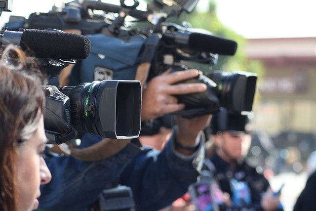 A group of camera operators holds various professional video cameras, ready to capture footage at an outdoor event.