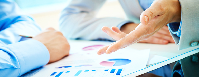 Two professionals discussing financial graphs and charts at a table, one gesturing with their hand while the other listens attentively.
