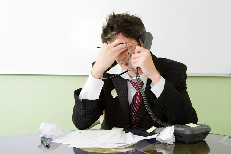 A frustrated businessman in a suit sits at a cluttered desk, holding a phone to his ear and crumpled papers scattered around him