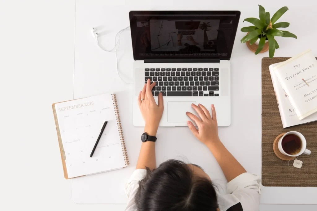 A woman with dark hair works on a laptop at a desk, with a calendar, pen, plant, and a tea cup nearby.