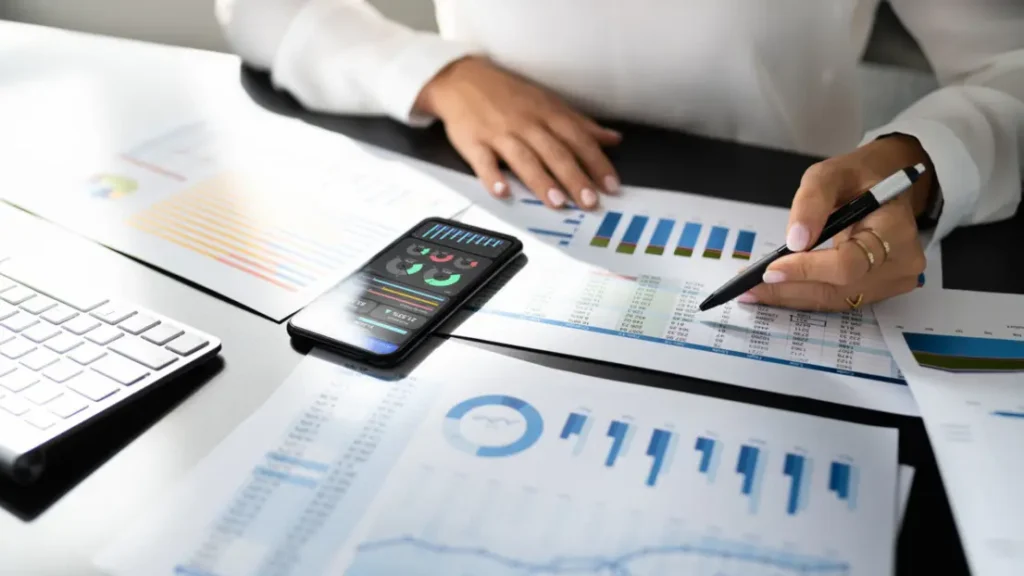 A person analyzing financial reports and graphs, using a smartphone for data, with a pen in hand and a keyboard nearby.