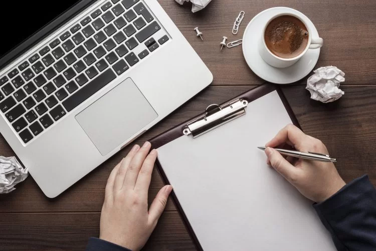 A hand holds a pen above a blank clipboard, beside a laptop and a cup of coffee, with crumpled paper scattered on a wooden desk.