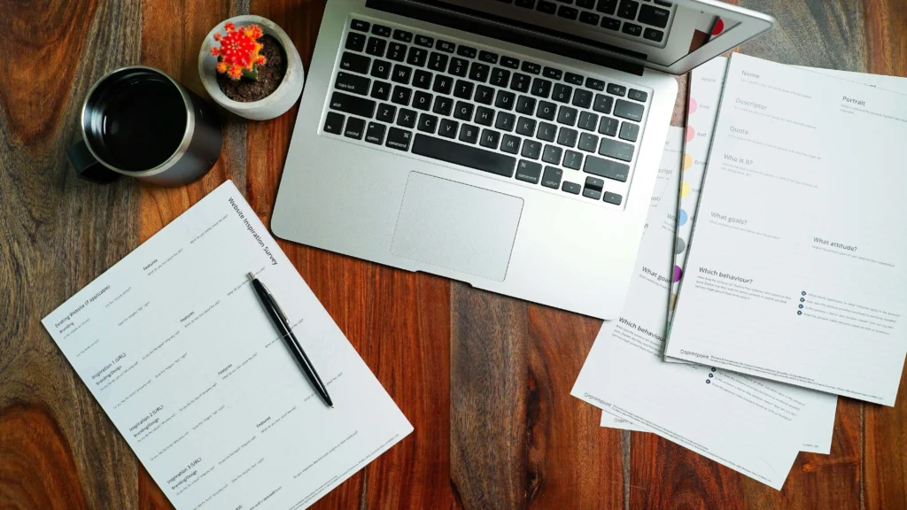 A laptop on a wooden desk beside a coffee mug, notes, and a small potted cactus, creating a workspace for planning and inspiration.