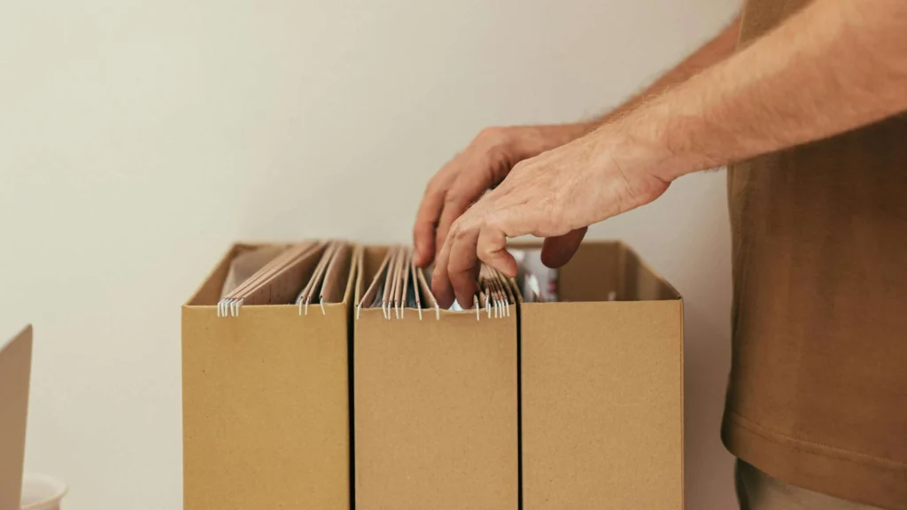 A person sorts through files in a brown cardboard box, organizing paperwork with their hands against a neutral background.
