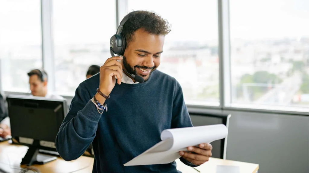 A person wearing headphones is holding a sheet of paper while sitting at a desk with a city view in the background.
