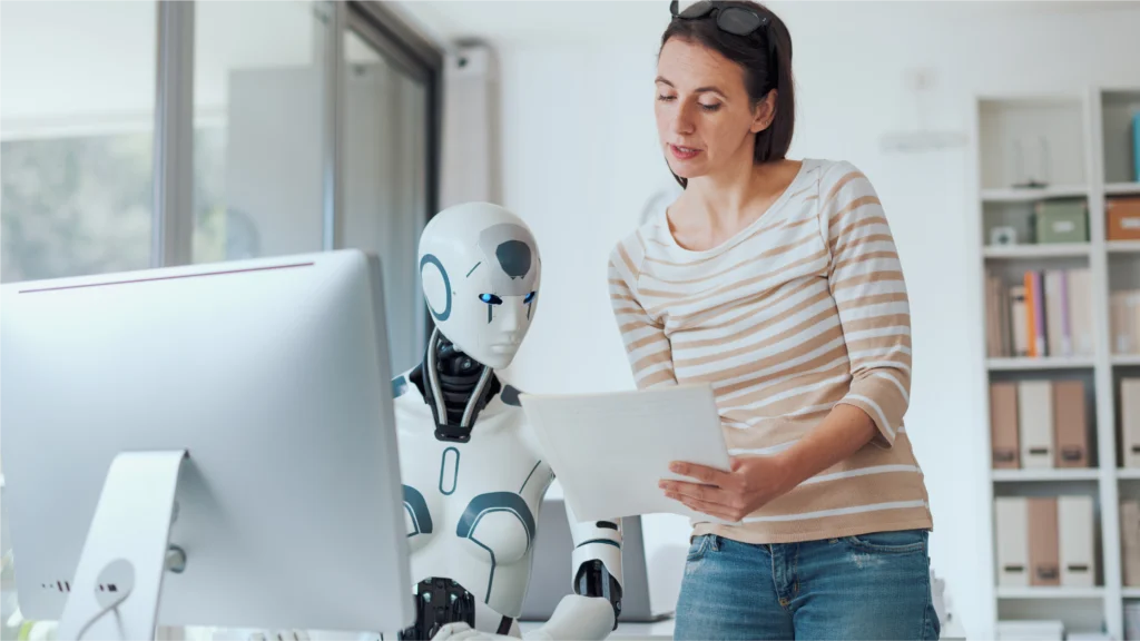 A woman in casual attire stands beside a humanoid robot, reviewing documents in a bright, modern office space with a desktop computer.