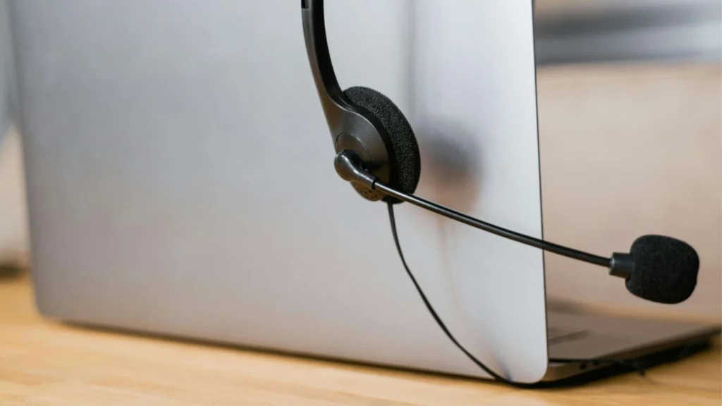 Close-up of a black headset with a microphone attached to the back of a laptop on a wooden table.