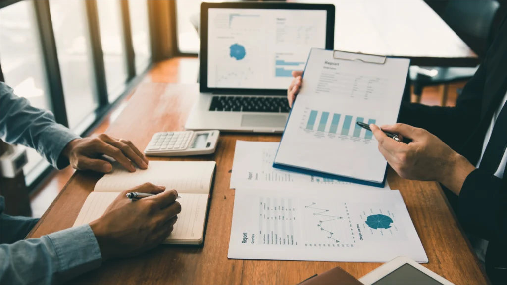 Two professionals discuss financial reports and data analysis at a wooden table, with a laptop, calculator, and notebooks present.