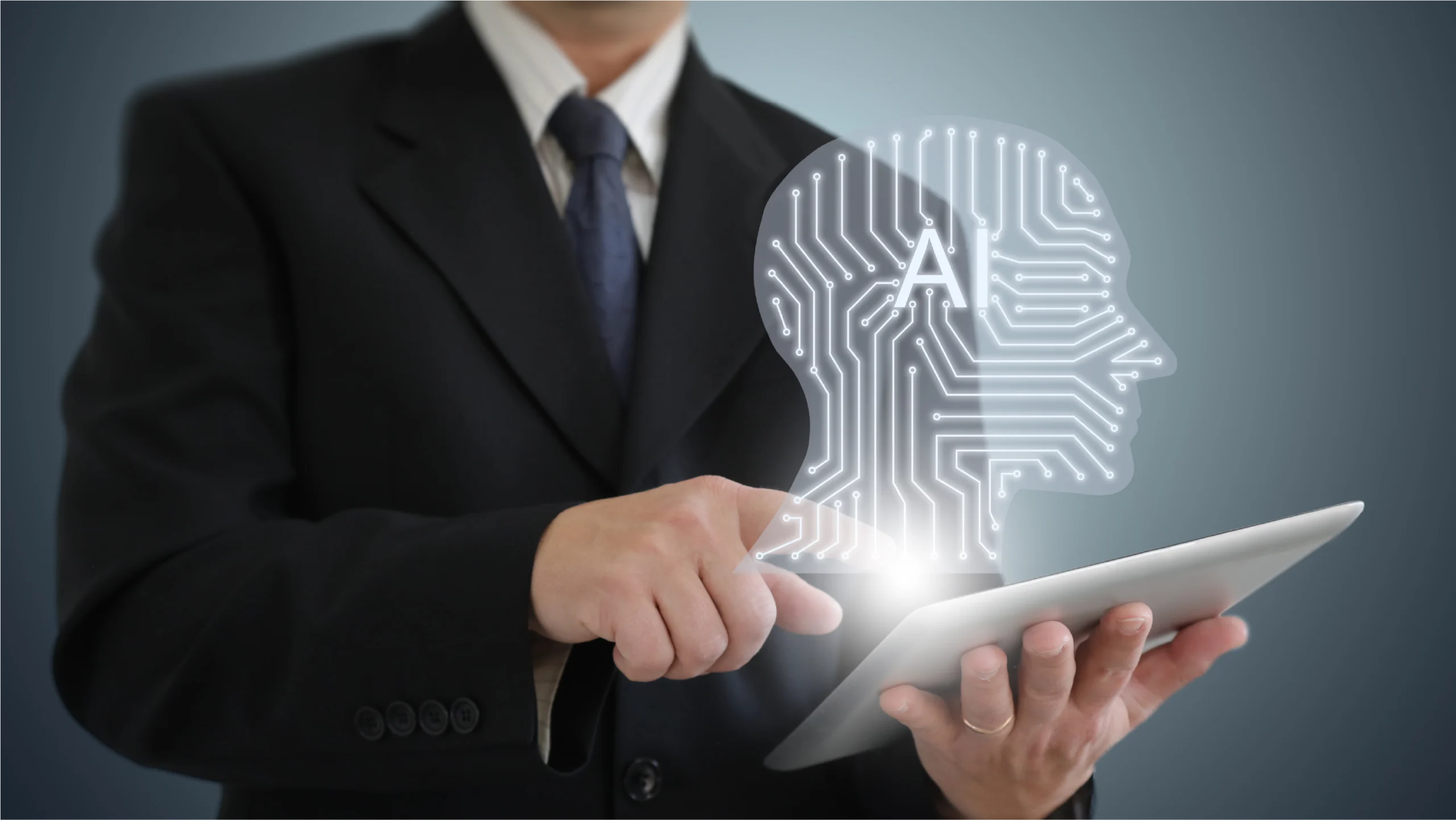 A businessman in a suit points at a tablet displaying a glowing human head silhouette with circuit patterns, symbolizing artificial intelligence.