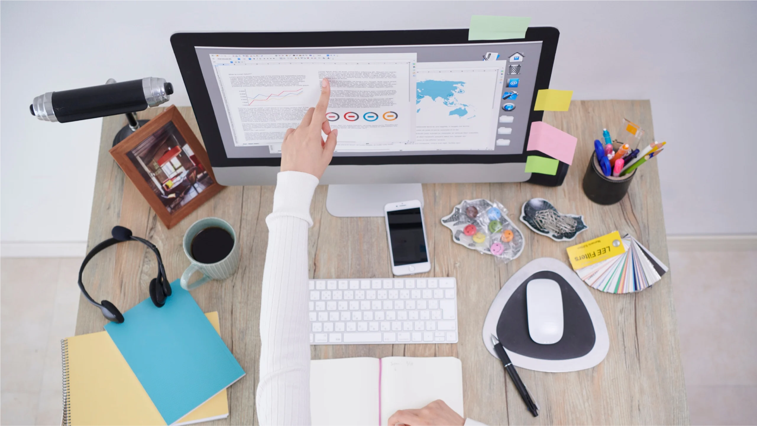 A person points at a document on a computer screen surrounded by a notebook, phone, coffee, and colorful office supplies on a wooden desk.