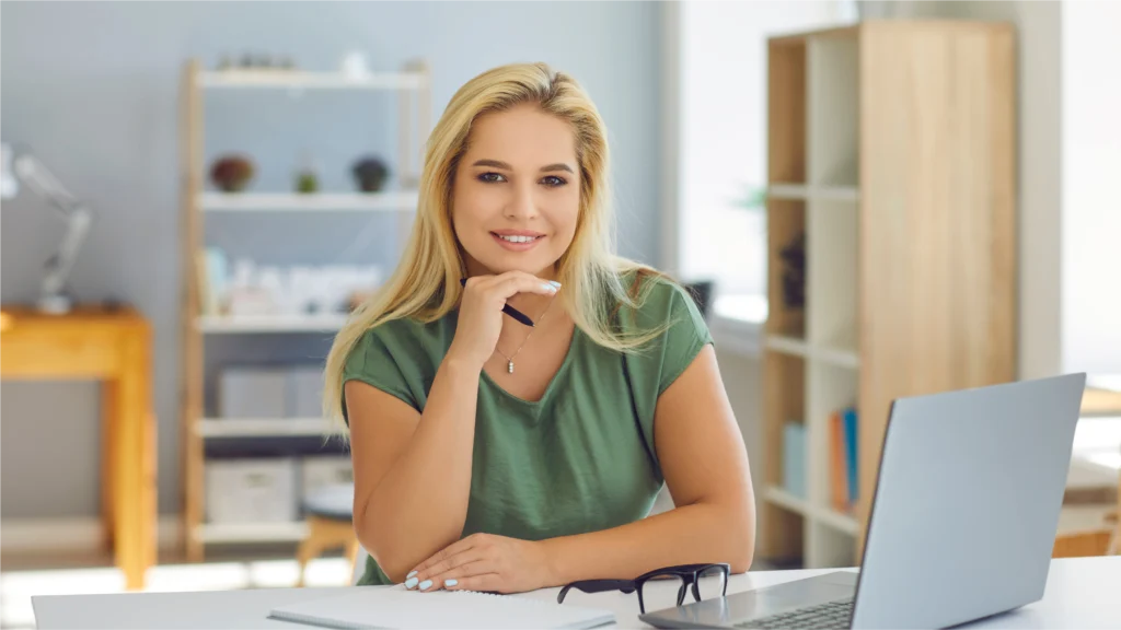 A woman with long blonde hair sits at a desk, holding a pen thoughtfully, with a laptop and glasses nearby in a well-lit room.