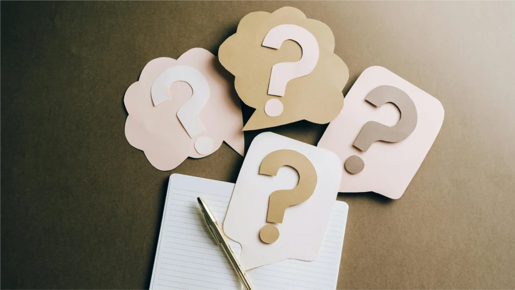 Colorful paper question marks surround a blank notebook and pen on a textured brown background, symbolizing inquiry and brainstorming.