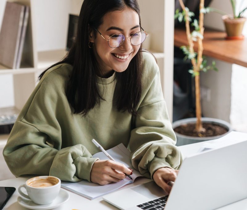 Smiling woman in glasses writing notes beside a laptop