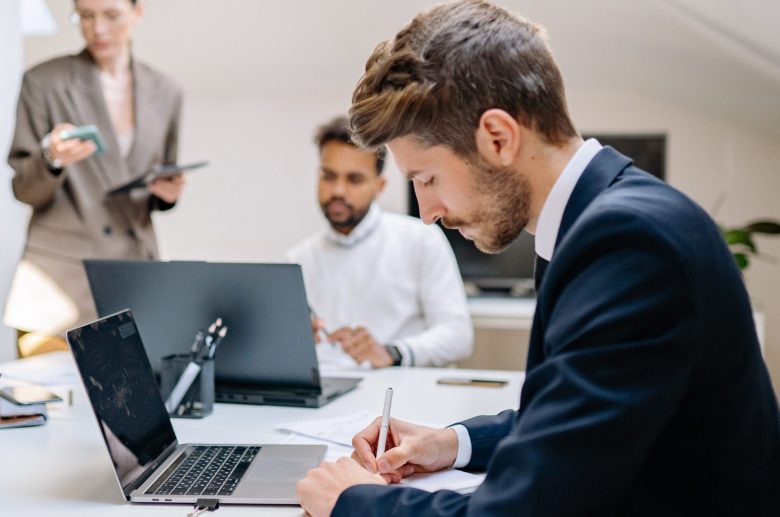 Man writing notes at a desk while colleagues work behind him