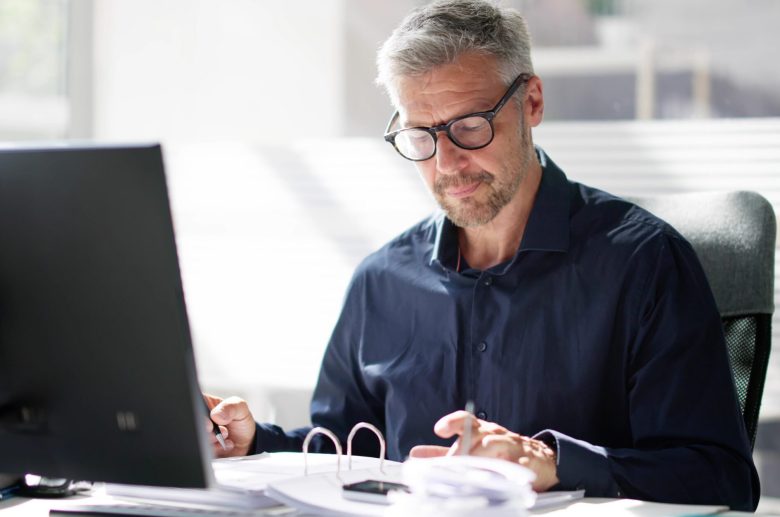 Man in glasses reviewing paperwork at a desk with a computer