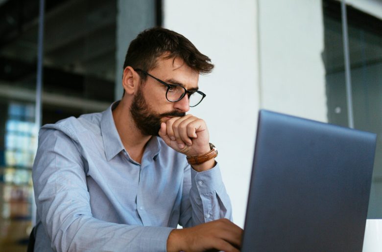 Man in glasses thinking while working on a laptop
