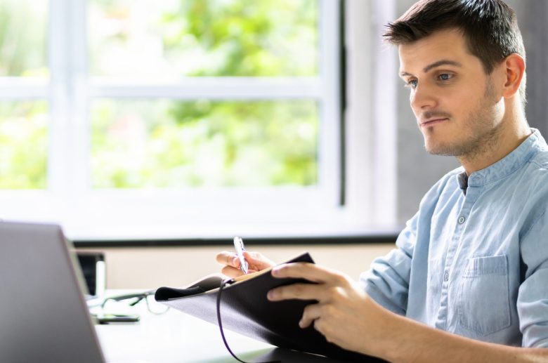 Man writing in a notebook next to a laptop