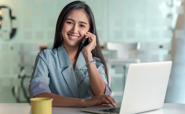 Asian Beautiful smiling businesswoman talking on the phone looking at the camera happy a coffee mug and laptop placed on the table.