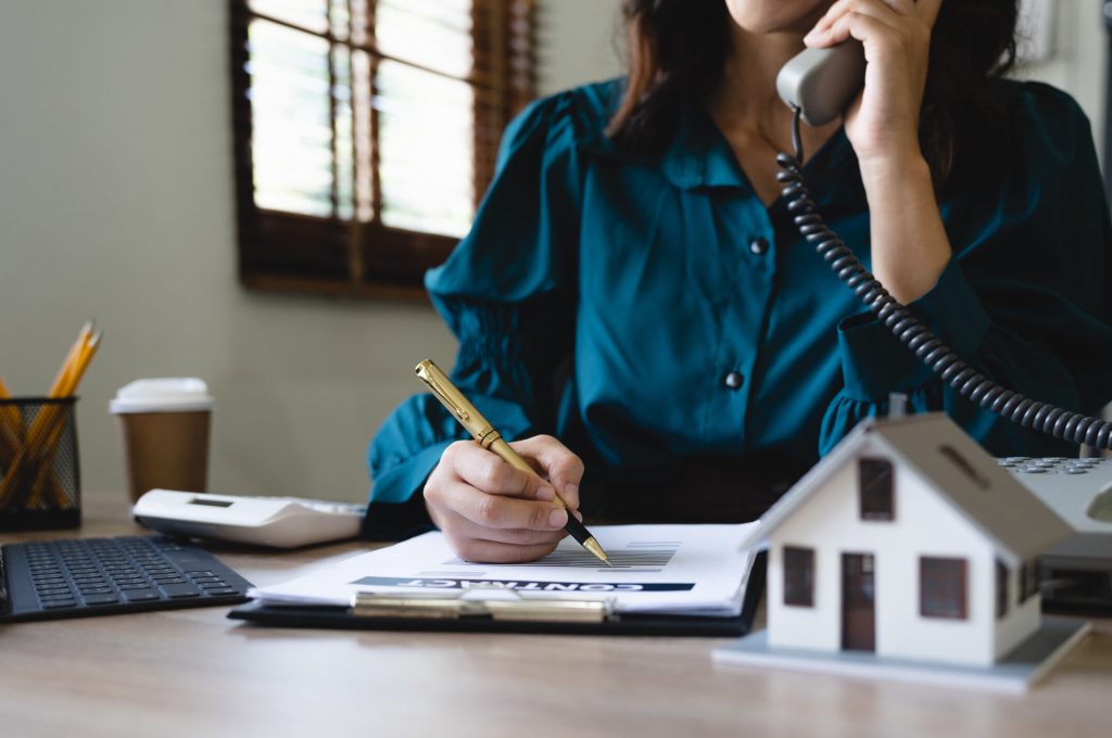 Woman on a desk phone taking notes, with a small house model on the desk