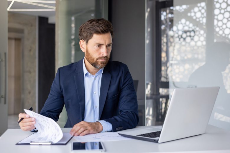 Man in a suit reviewing papers at a desk with a laptop