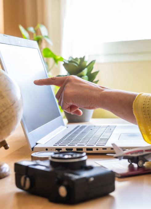 Woman pointing at laptop screen while planning a trip on a booking website, surrounded by a globe, camera, passport, and airplane model on the desk, creating an organized itinerary